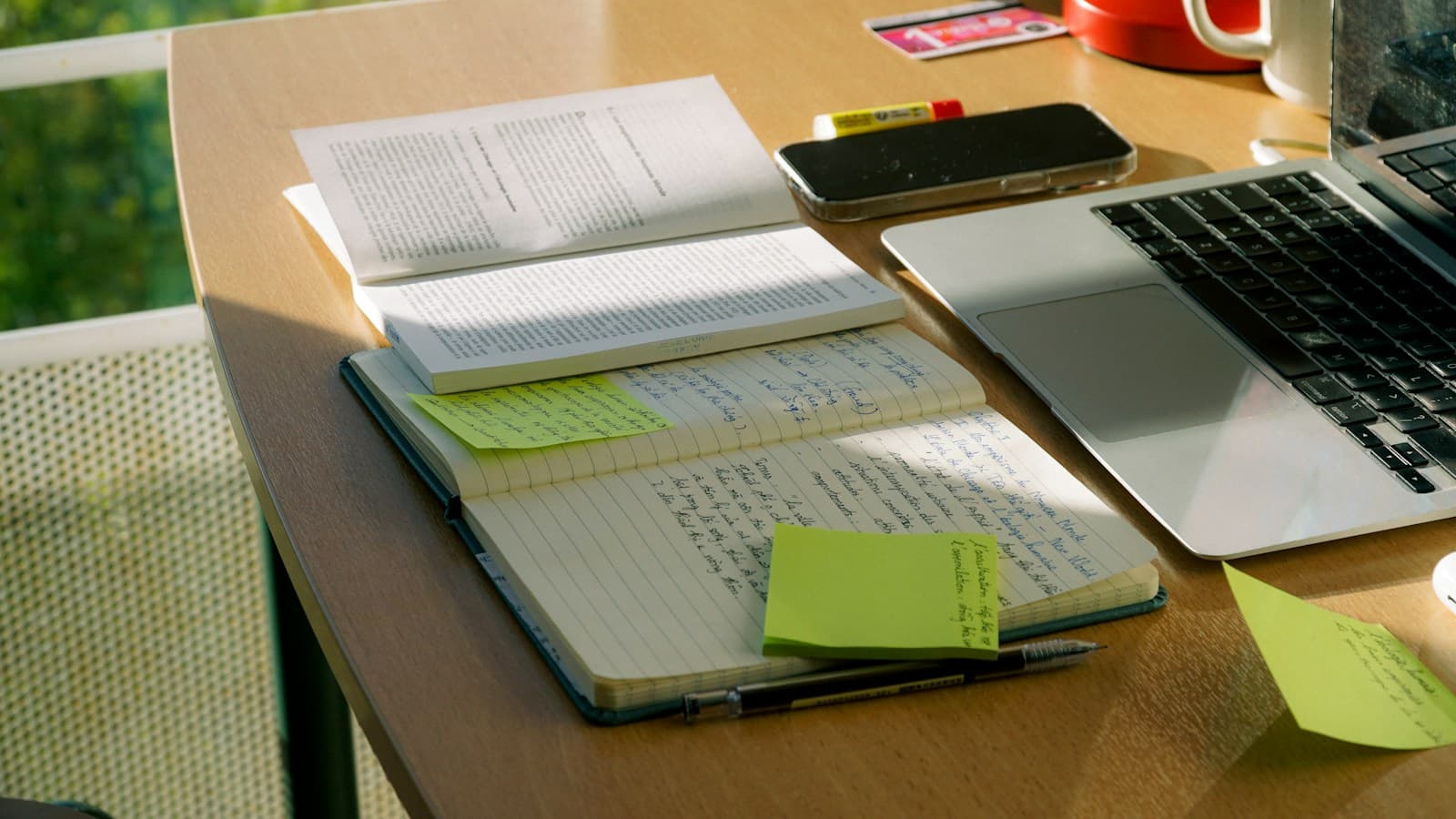 A sunlit desk with a laptop, open notebook, and handwritten notes